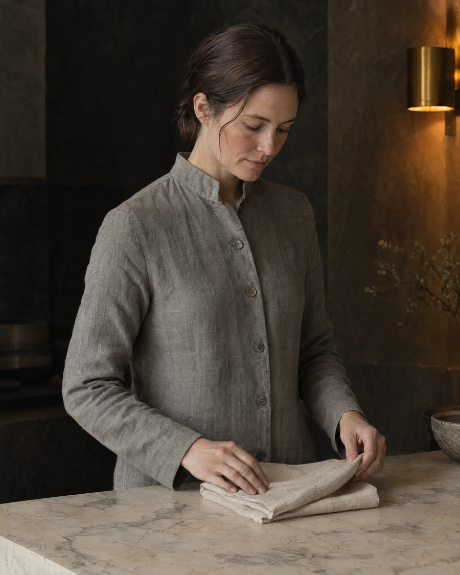 A practitioner in a stone grey linen coat folds a square of undyed linen at a honed marble counter.